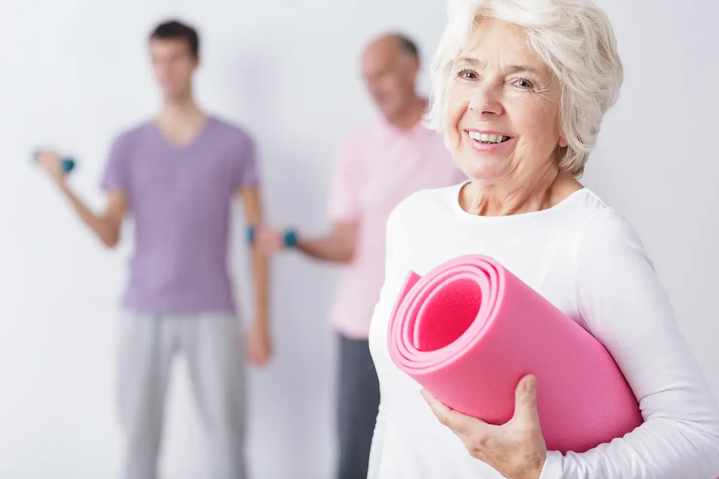 An older lady smiles as she holds a rolled up yoga mat. Two other people are out of focus behind her.